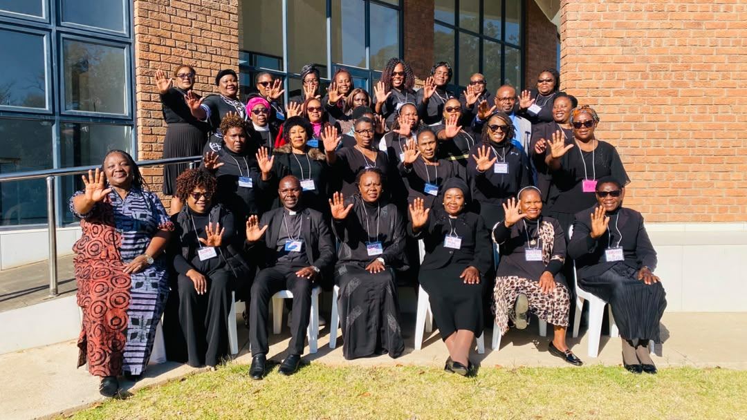 Participants dressed in black raise a hand each for the camera, seated and standing outside.