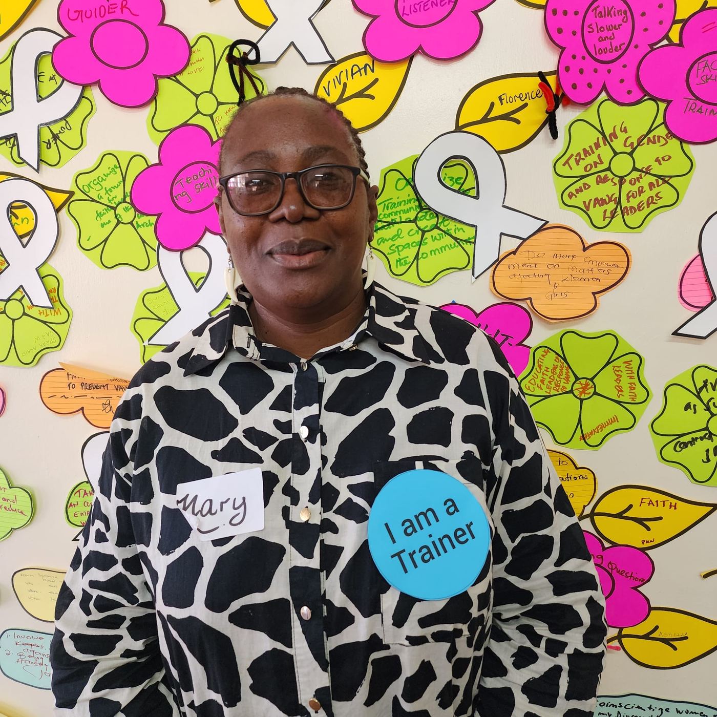 A participant wearing an I Am A Trainer badge stands in front of the wall of paper leaves and flowers.